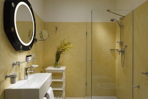 A bathroom with a large mirror, a white sink, a glass shower enclosure, and a vase with yellow flowers on a small shelf.