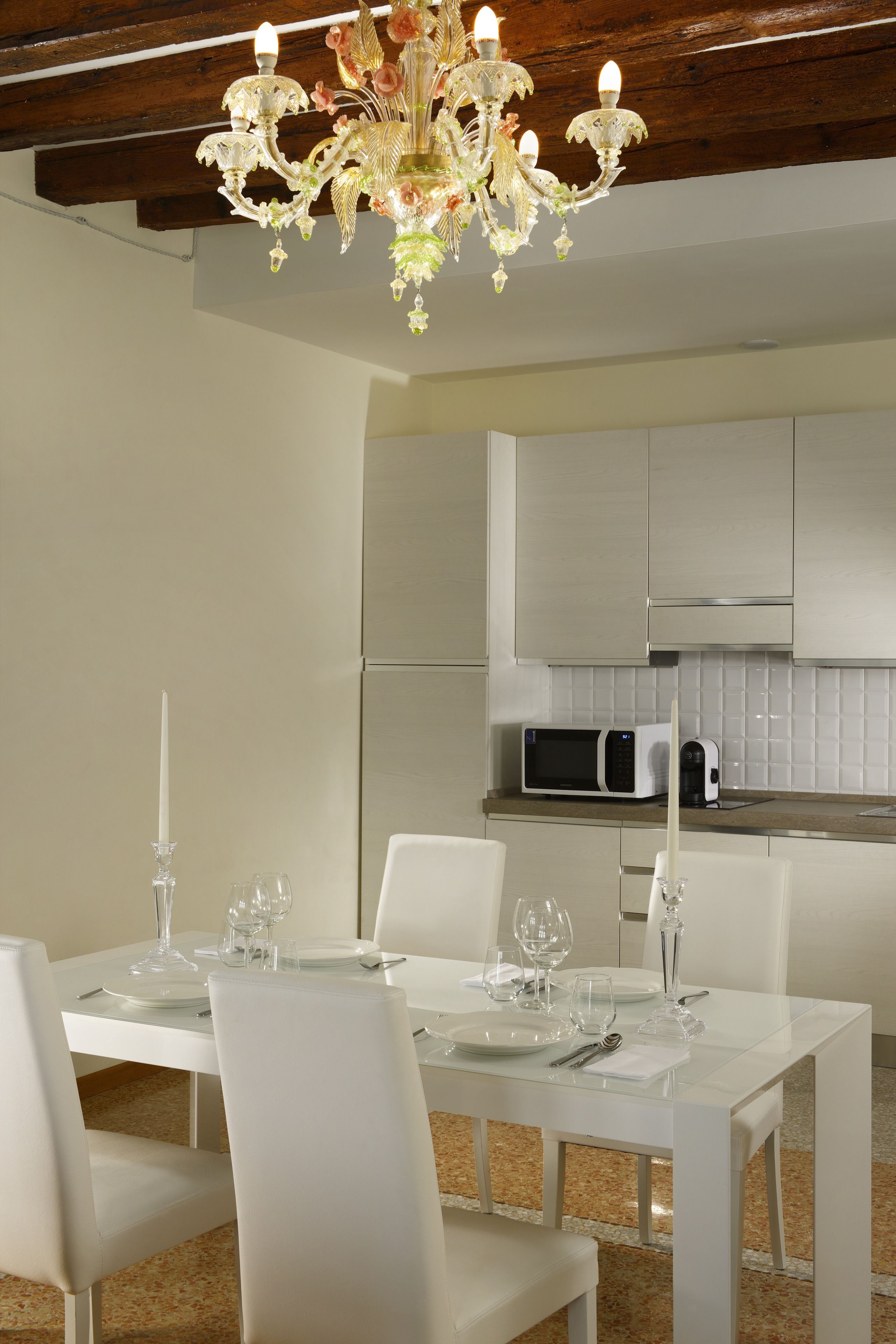 Un comedor con una mesa blanca dispuesta para cuatro personas, una araña y una cocina al fondo.