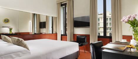 A hotel room with a large bed, a desk with a chair, a flat-screen TV, and a view of buildings through the window.