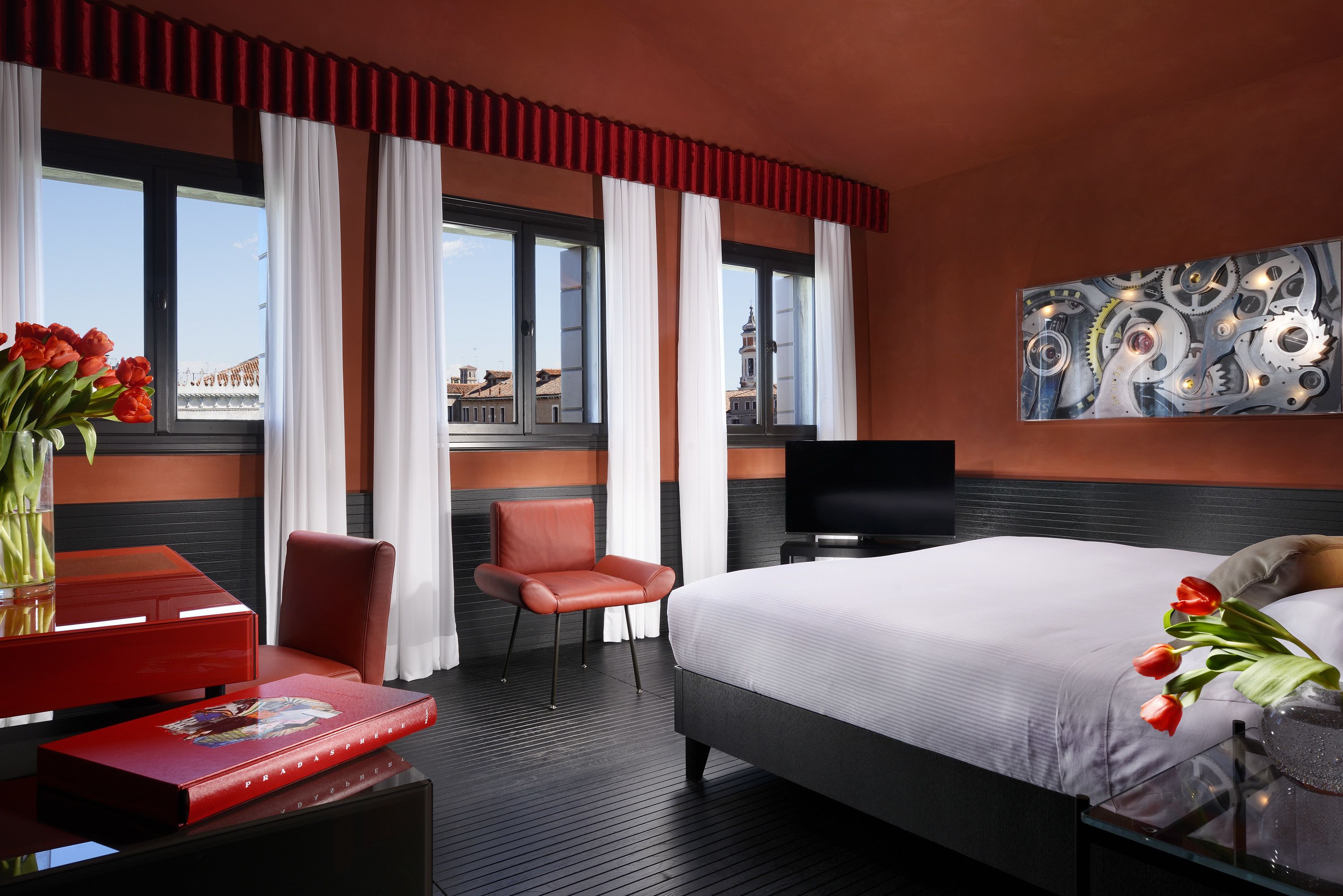 A modern hotel room with a large bed, a red leather chair, a desk with a red book, and a painting on the wall.