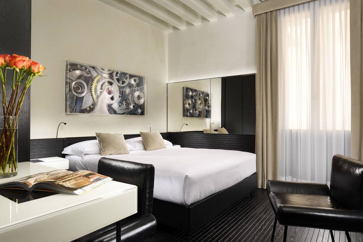 A modern hotel room with a large bed, a desk, and a chair. There are abstract paintings on the walls and a large window with curtains.