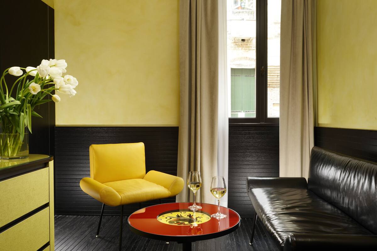 A yellow armchair and a black leather sofa in a room with a round table between them, a vase with white flowers, and a window with curtains.