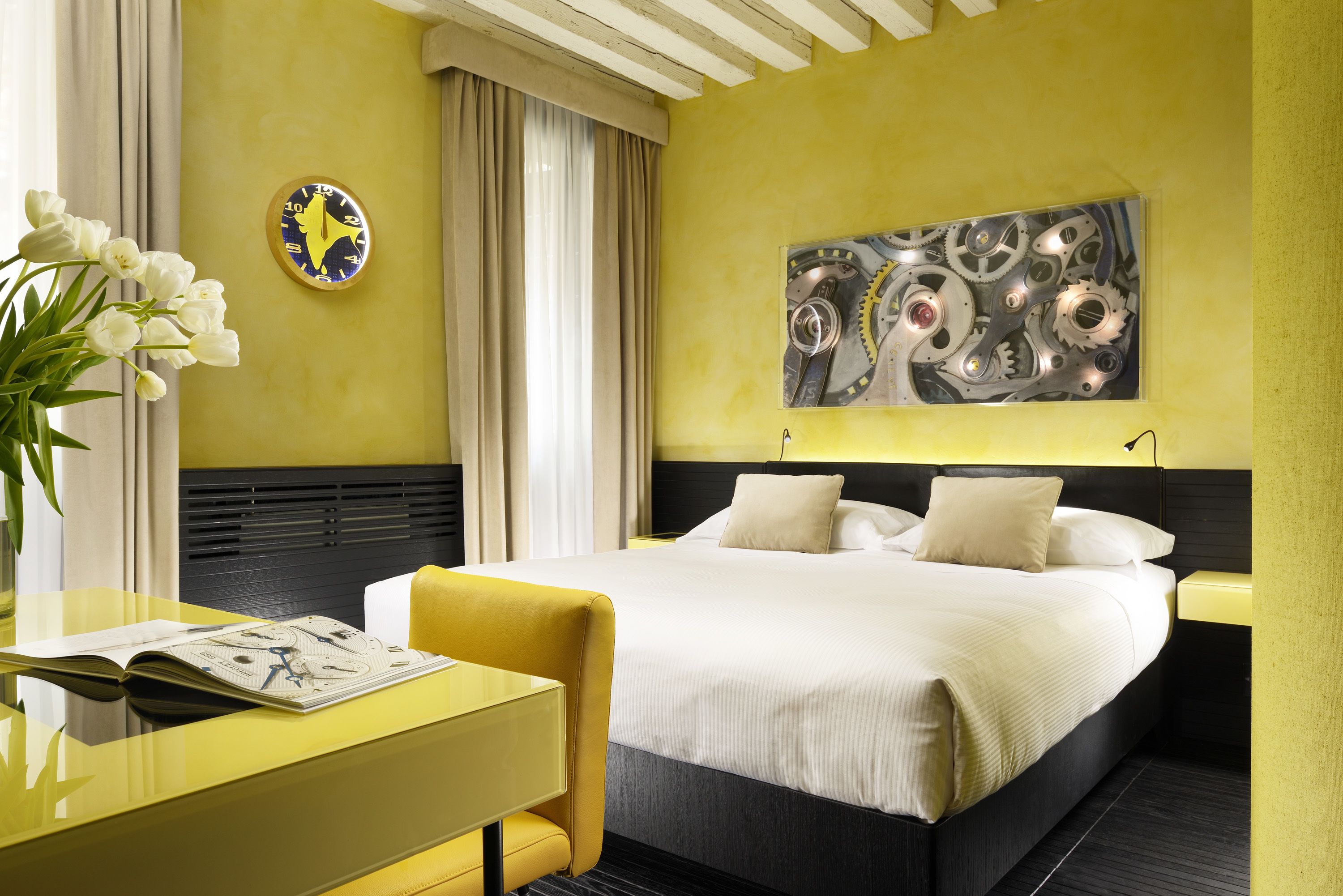 Quarto de hotel com cama, mesa de cabeceira, uma cadeira, luminária, relógio e um quadro na parede.