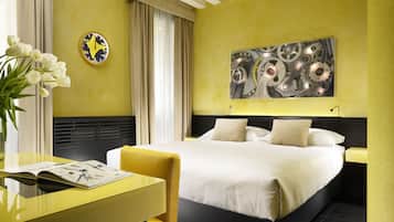 Quarto de hotel com cama, mesa de cabeceira, uma cadeira, luminária, relógio e um quadro na parede.