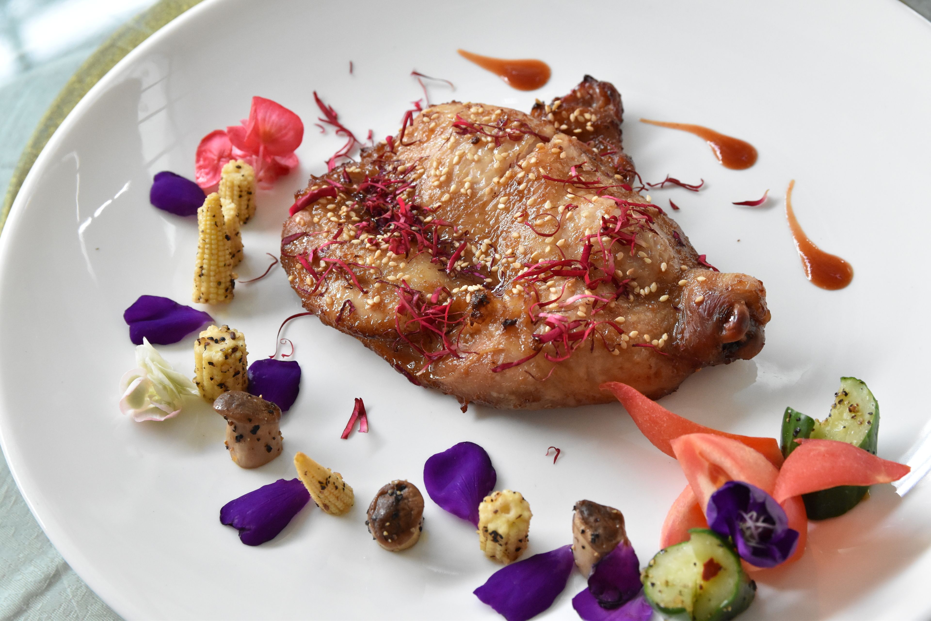 Une assiette contenant un plat de viande cuite, garni de graines de sésame et de pétales de fleurs rouges, accompagné de légumes et d’une sauce.