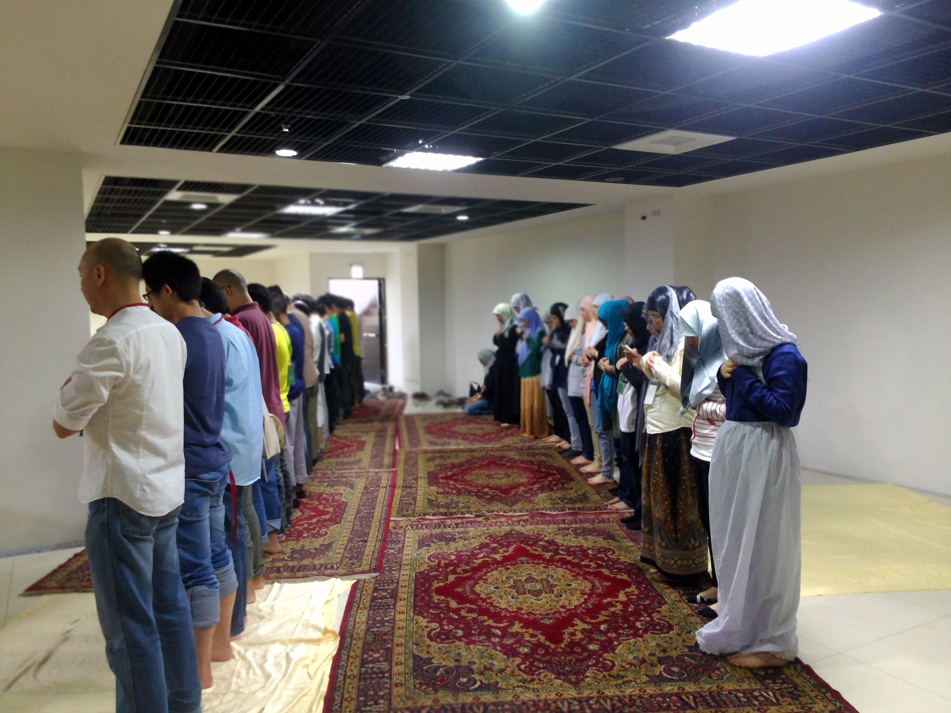 A prayer hall with a patterned carpet, a high ceiling with grid-like panels, and a group of people standing in a line.