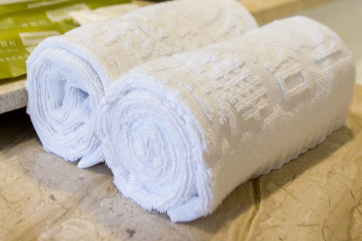 Two rolled white towels on a wooden surface, a green bottle, and a brown vase with a plant in a bathroom setting.