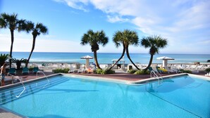 A swimming pool with clear blue water, surrounded by palm trees and lounge chairs, with a view of the ocean in the background.