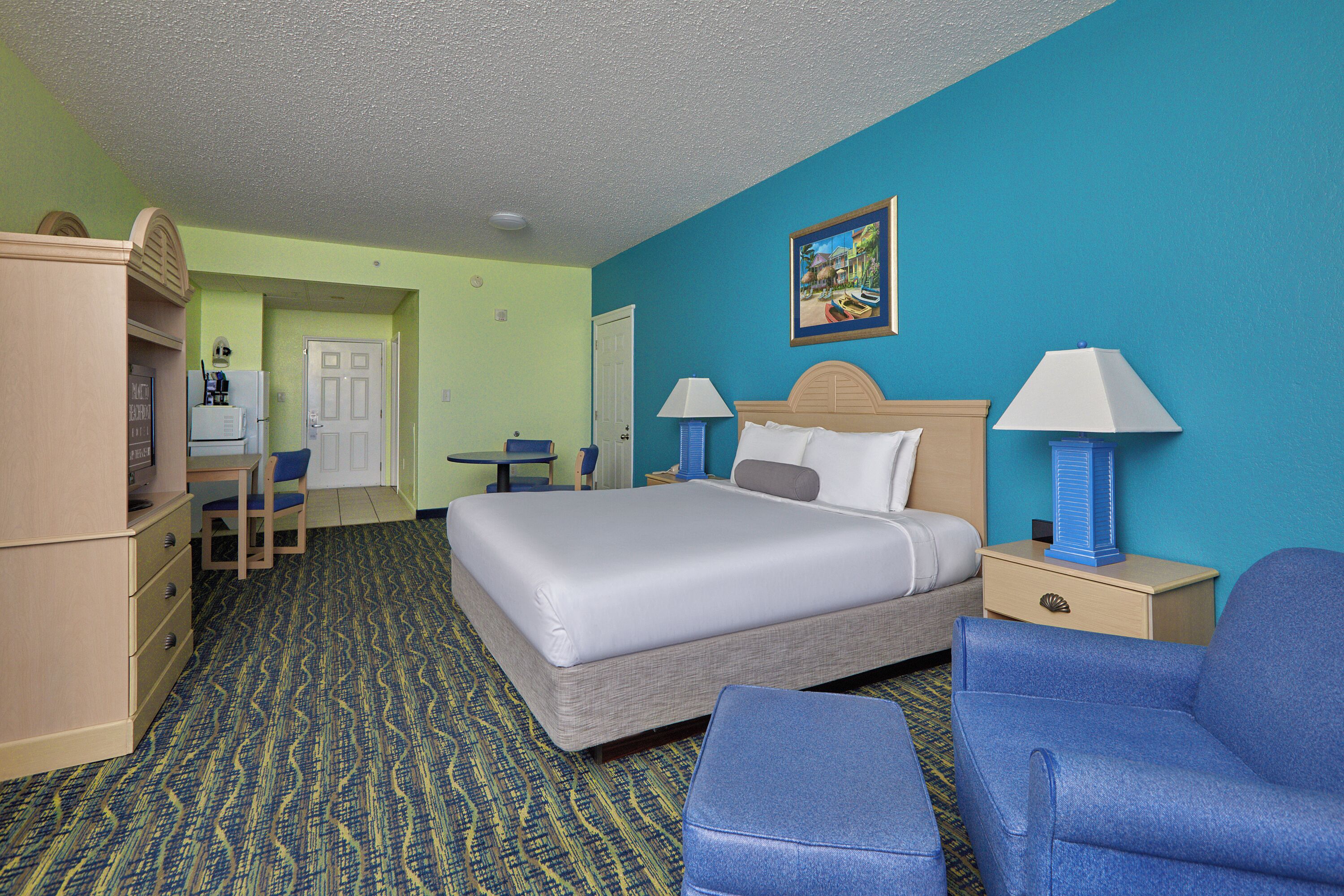 A hotel room with a bed, two chairs, a desk, a dresser, and a painting on the wall.