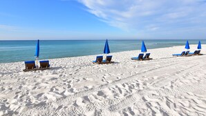 A beach with white sand and clear blue water. There are several blue umbrellas and wooden lounge chairs arranged in rows.