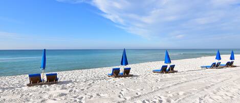 Une plage de sable blanc bordée d’une eau d’un bleu cristallin. On y trouve plusieurs parasols bleus et des chaises longues en bois disposées en rangées.