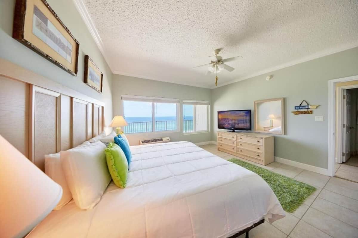 A bedroom with a bed, a television, a ceiling fan, a dresser, and a view of the ocean.
