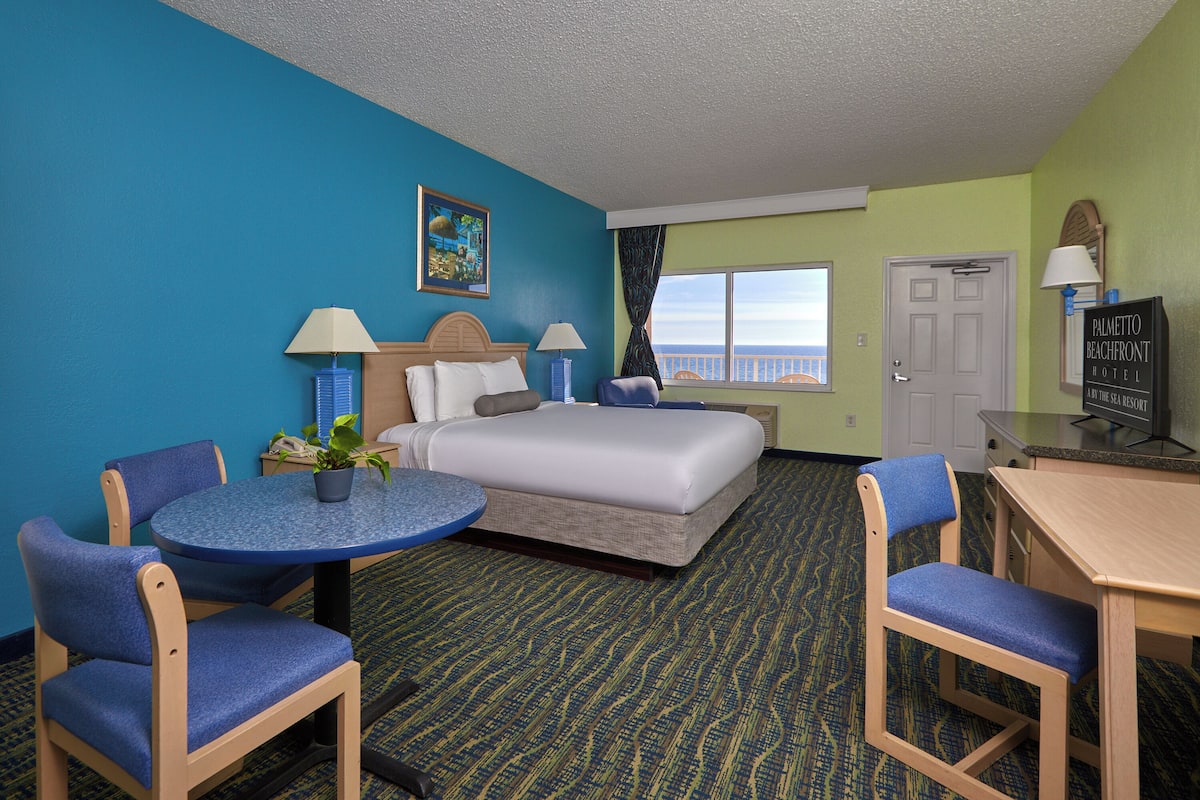 A hotel room with a bed, two chairs, a small table, a desk, a television, and a view of the ocean.