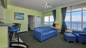 A room with a sofa, chairs, a small table, a ceiling fan, a view of the ocean, and a door leading to another room.