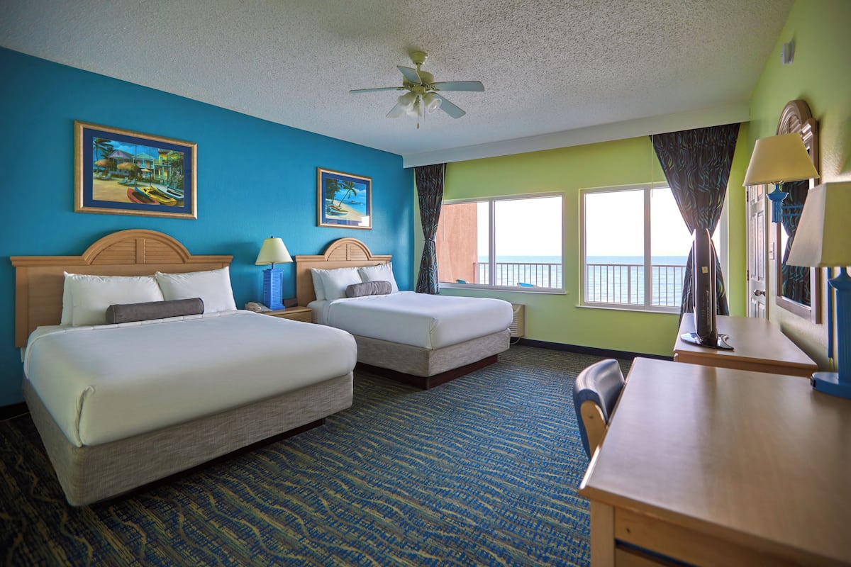 A hotel room with two beds, a desk, a chair, a ceiling fan, and large windows with curtains.