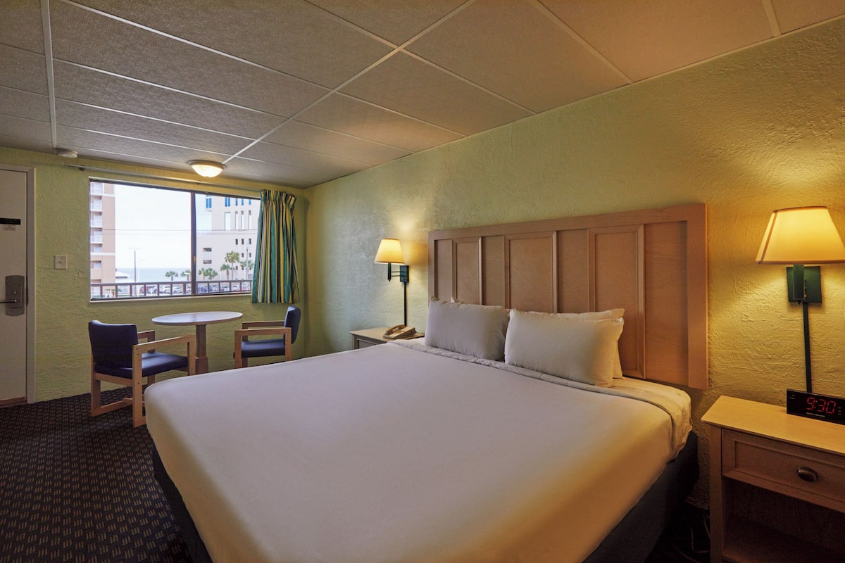 A hotel room with a large bed, a desk with a lamp, a chair, a small table, and a window with curtains.