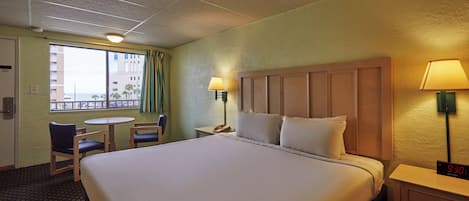 Une chambre d’hôtel avec un grand lit, un bureau avec une lampe, une chaise, une petite table et une fenêtre avec des rideaux.