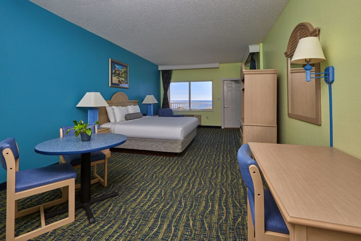 A hotel room with a bed, a desk, a chair, a small table, a lamp, a plant, and a view of the ocean.