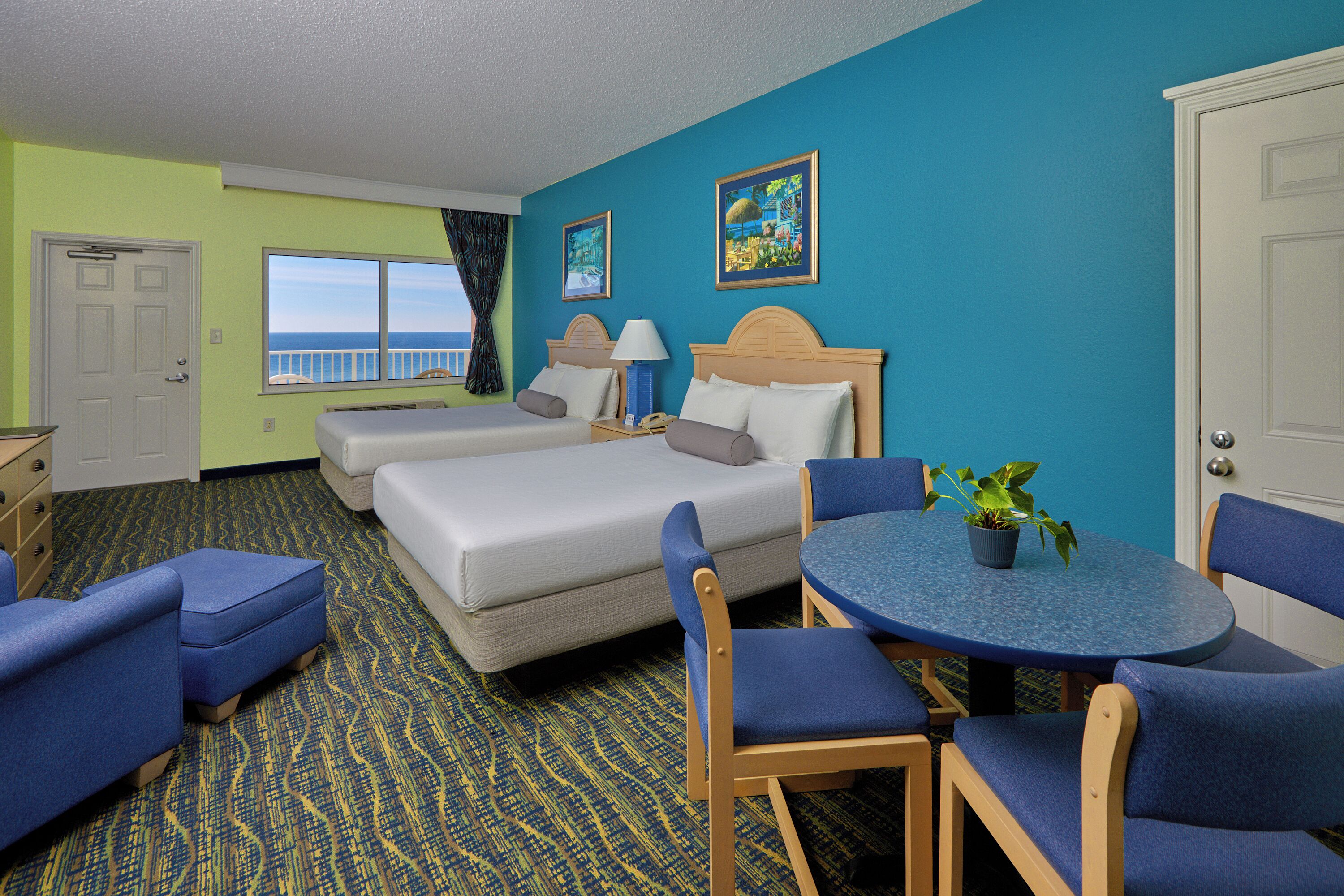 A hotel room with a bed, two chairs, a small table with a plant, a window with a view, and a door.