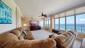 A spacious living room with a large brown sofa, a television, and a view of the ocean through large windows.