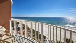 A balcony with a view of the beach, white chairs, and a table.