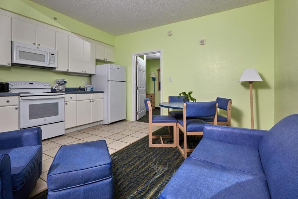 A compact kitchen with white appliances, a dining area with blue chairs, and a green wall.