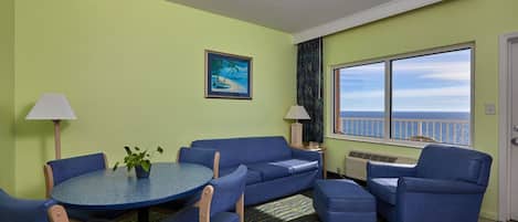 A room with a round table, blue sofa, and chairs, a window with a view of the sea, and a painting on the wall.