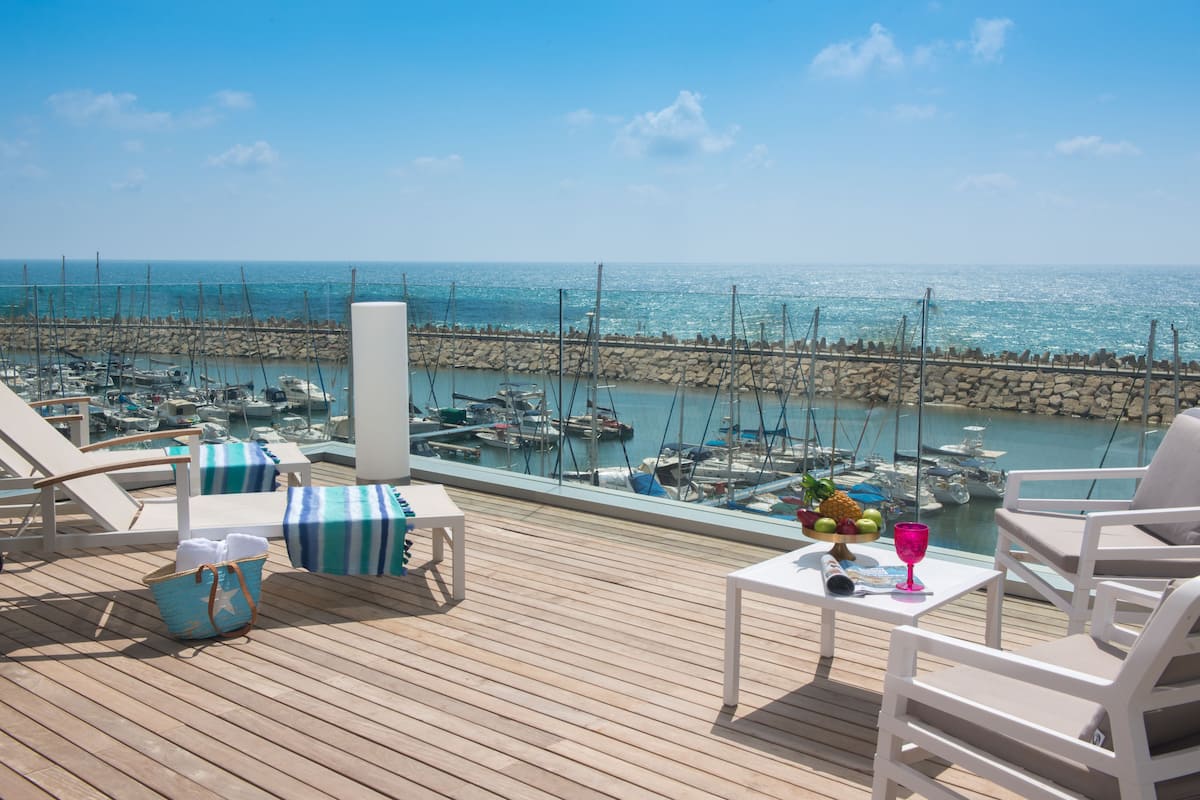 Een houten terras met lounge fauteuils en een tafel, met uitzicht op een jachthaven met boten en een stenen muur.