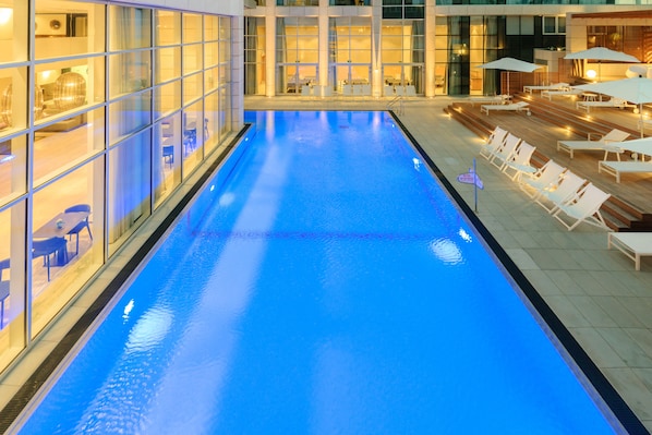 Une piscine entourée de chaises longues et de parasols, avec des murs en verre et des espaces de détente intérieurs.