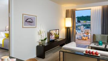 Habitación de hotel con cama, televisor, zona de comedor con mesa y sillas, balcón con vistas al paisaje y una planta decorativa.