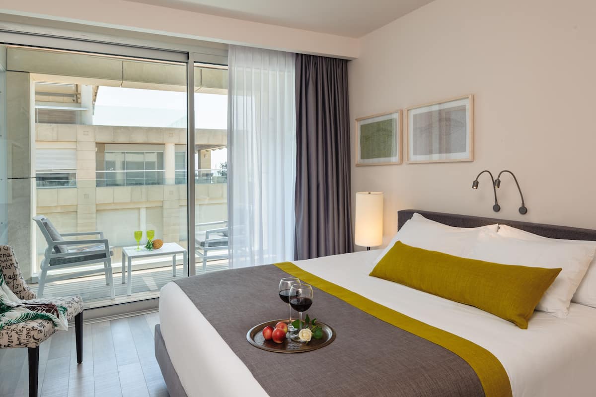Een hotelkamer met een bed, een eettafel met fruit en wijn, een stoel, en een balkon met uitzicht op gebouwen.