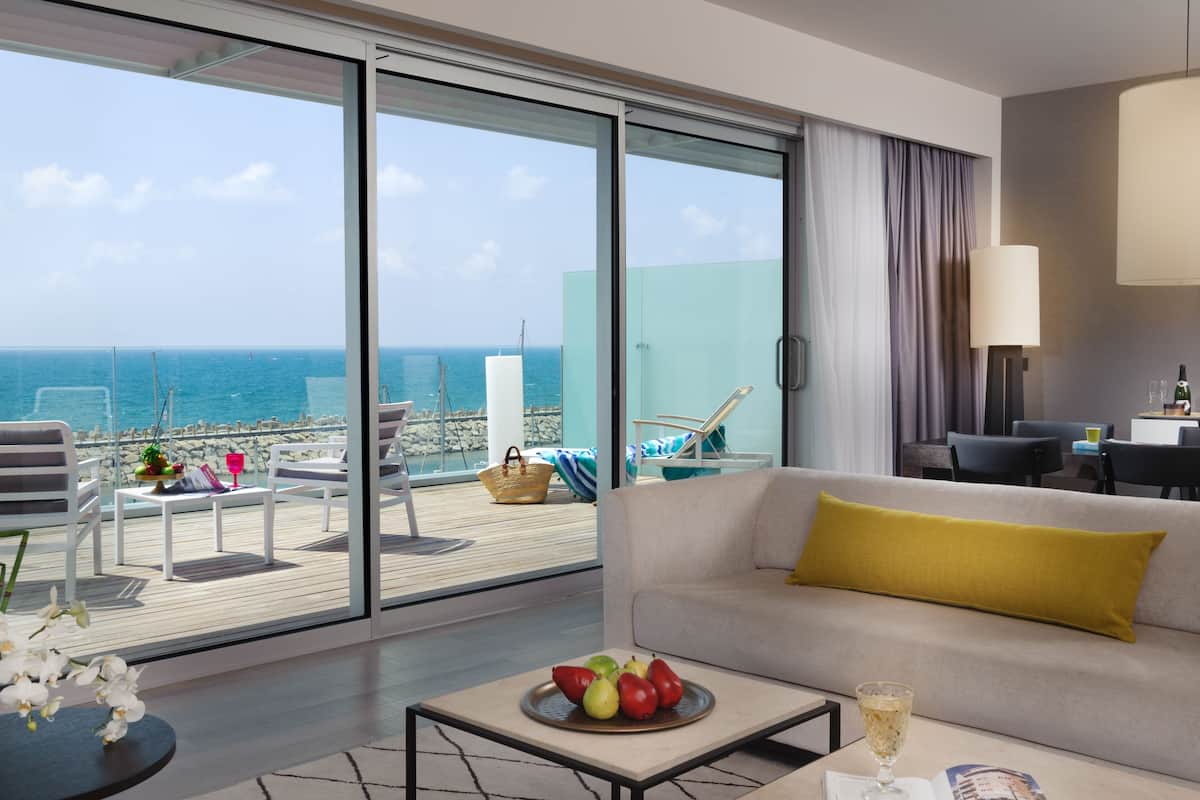 Een moderne hotelkamer met een grote schuifglazen deur die leidt naar een balkon met uitzicht op zee. Er is een bank met een geel kussen, een bijzettafeltje met fruit en een glas champagne, en een eethoek met stoelen en een tafel.