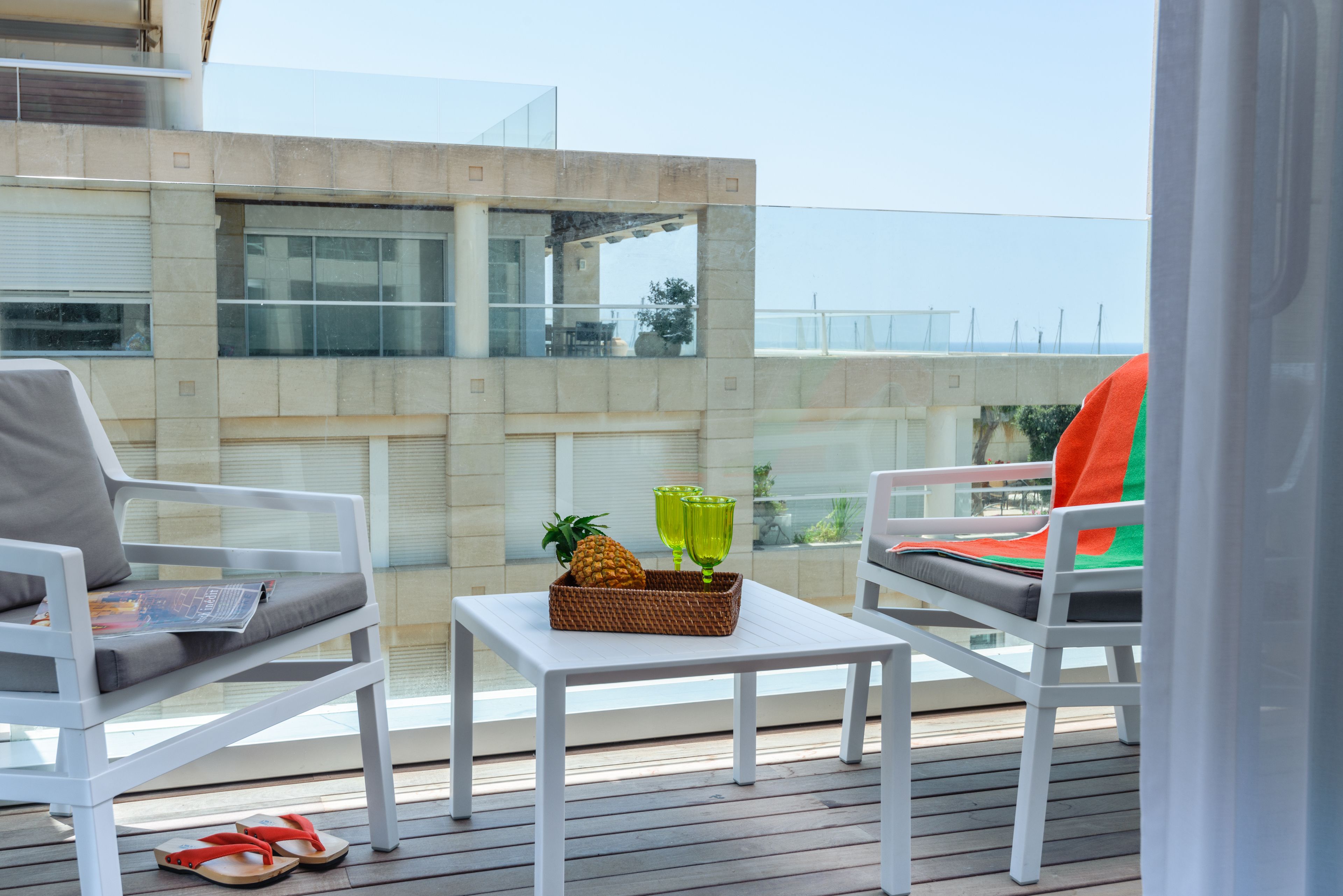 A balcony with white chairs, a small table, and a basket containing a pineapple and green glasses.