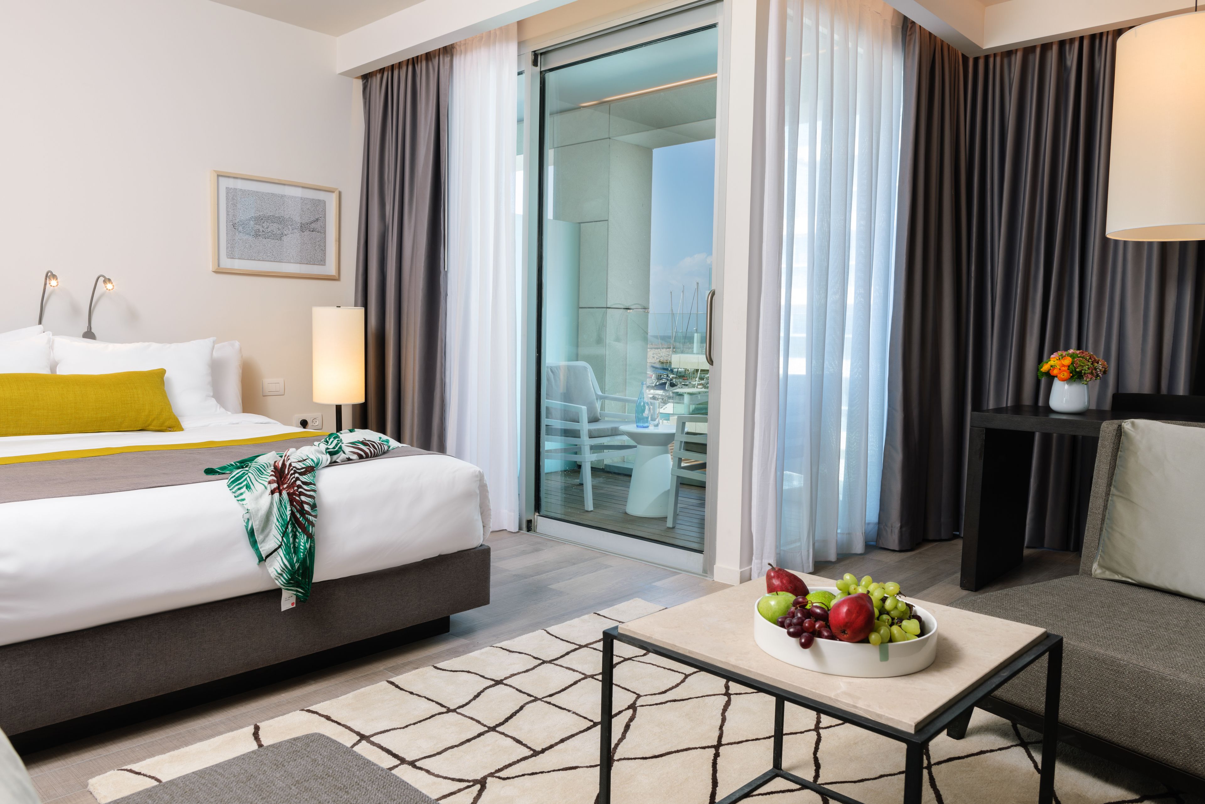 A hotel room with a bed, a dining area with a table and chairs, a fruit bowl on a table, and a view through a glass door.