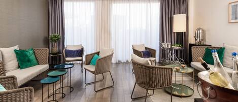 A modern living room with a sofa, two armchairs, a coffee table, and a dining table. There is a large window with curtains, a lamp, and a minibar.