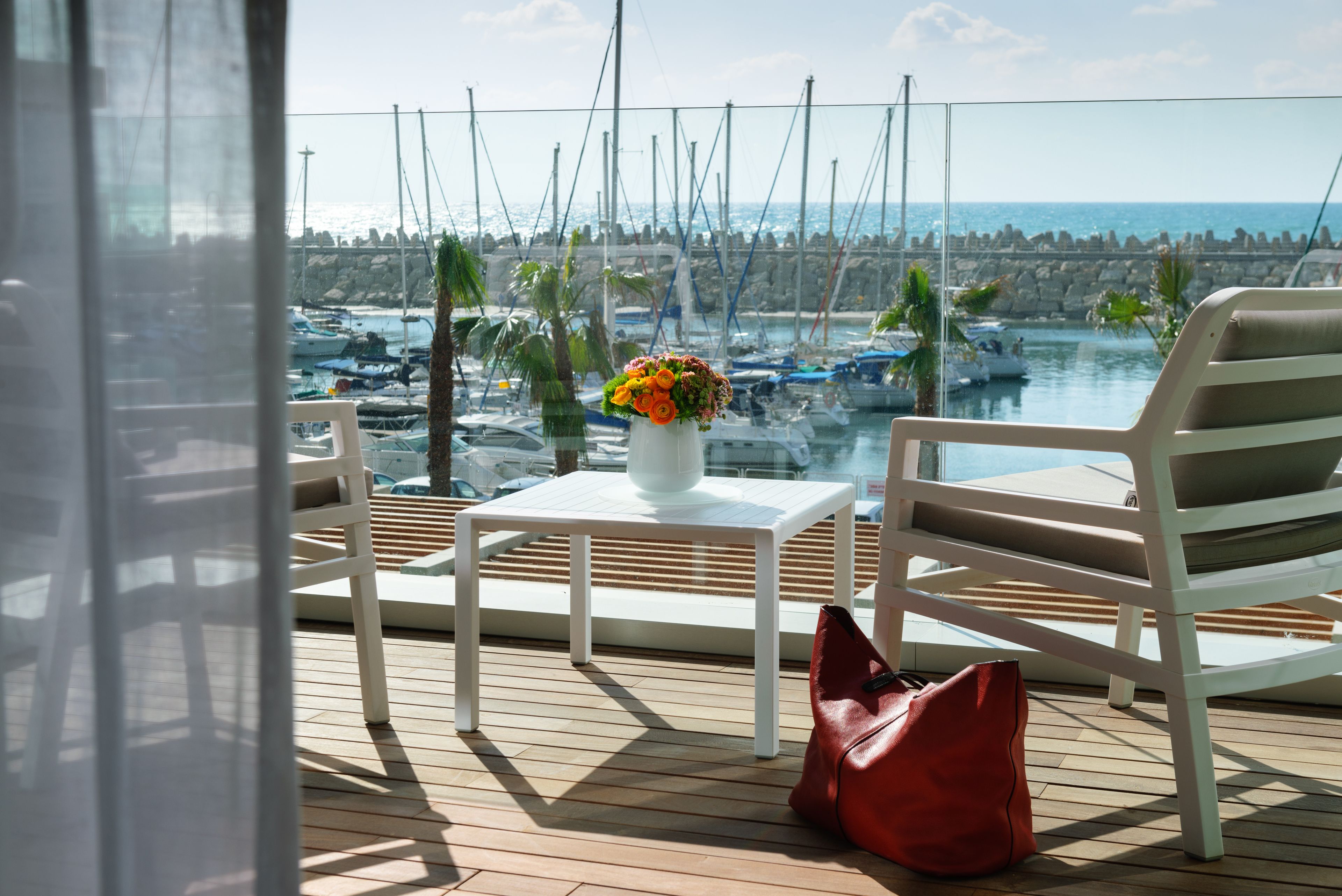 A balcony with a white table, a red bag, and a chair overlooking a marina with boats and a rocky shoreline.