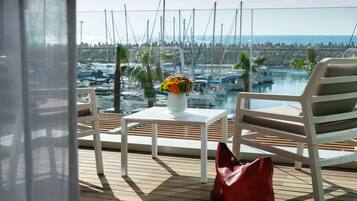 A balcony with a white table, a red bag, and a chair overlooking a marina with boats and a rocky shoreline.