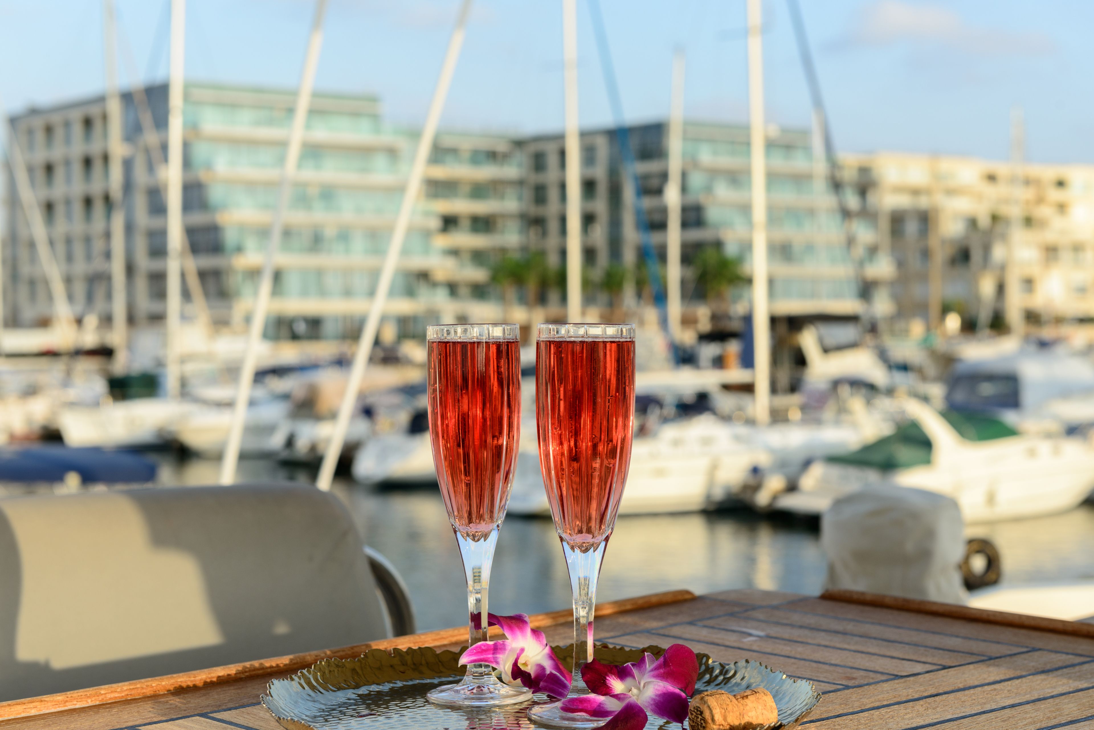 Two champagne flutes on a wooden tray with a flower, set against a marina with boats and modern buildings in the background.