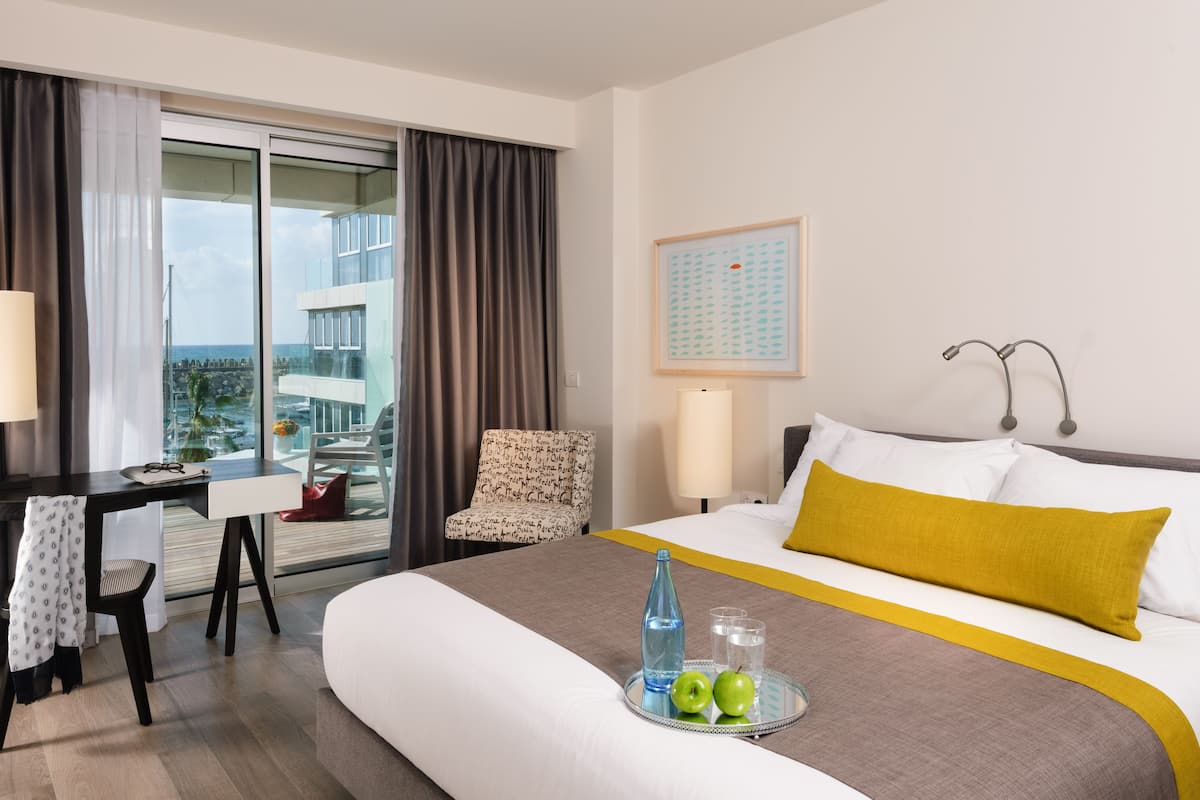 Hotelkamer met een bed, bureau, stoel, raam met gordijnen, een ingelijst beeld aan de muur en een dienblad met appels en glazen op het bed.