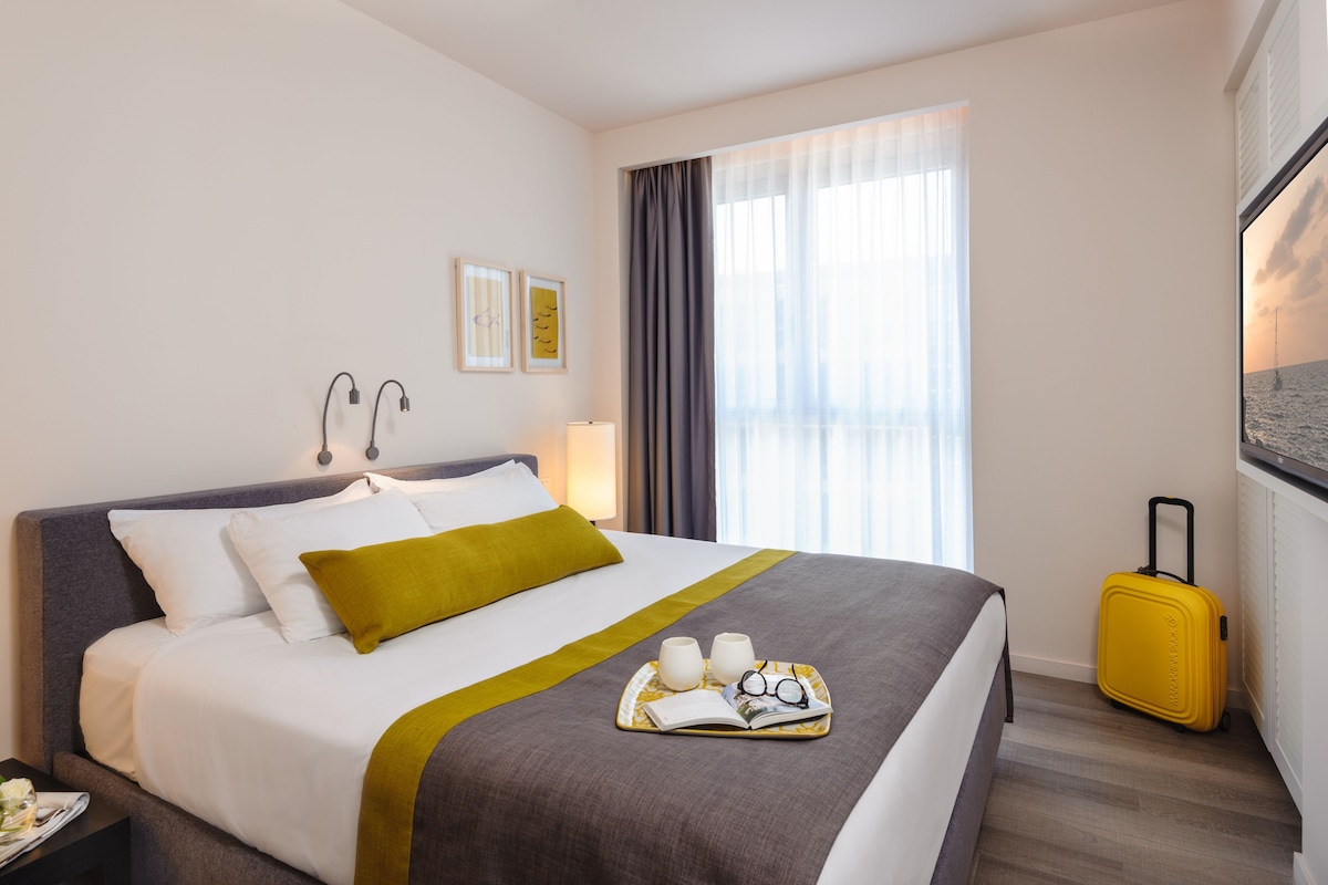 Habitación de hotel impecable con una cama grande, una mesita de noche con lámpara, una ventana con cortinas y una maleta.