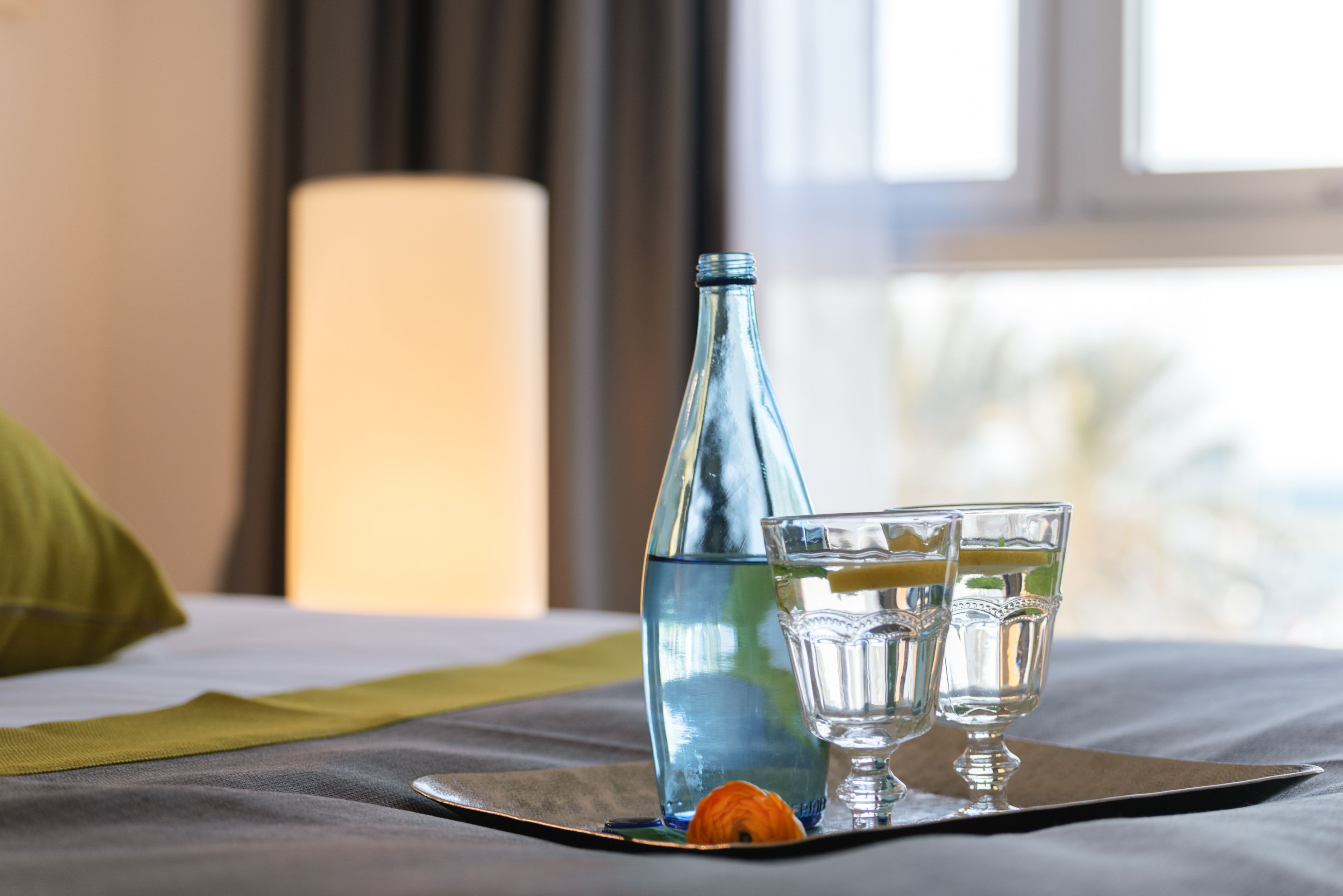 A bed with a tray containing a bottle of water, two glasses, and a lemon slice.