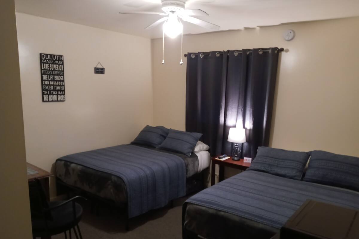 A hotel room with two single beds, a desk with a chair, a ceiling fan, and a sign listing local attractions.