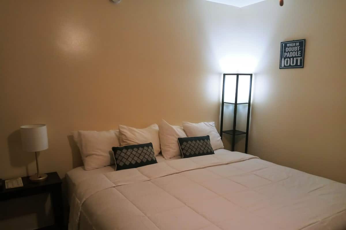 A neatly made bed with white bedding and two decorative pillows, a bedside lamp, and a wall-mounted sign.