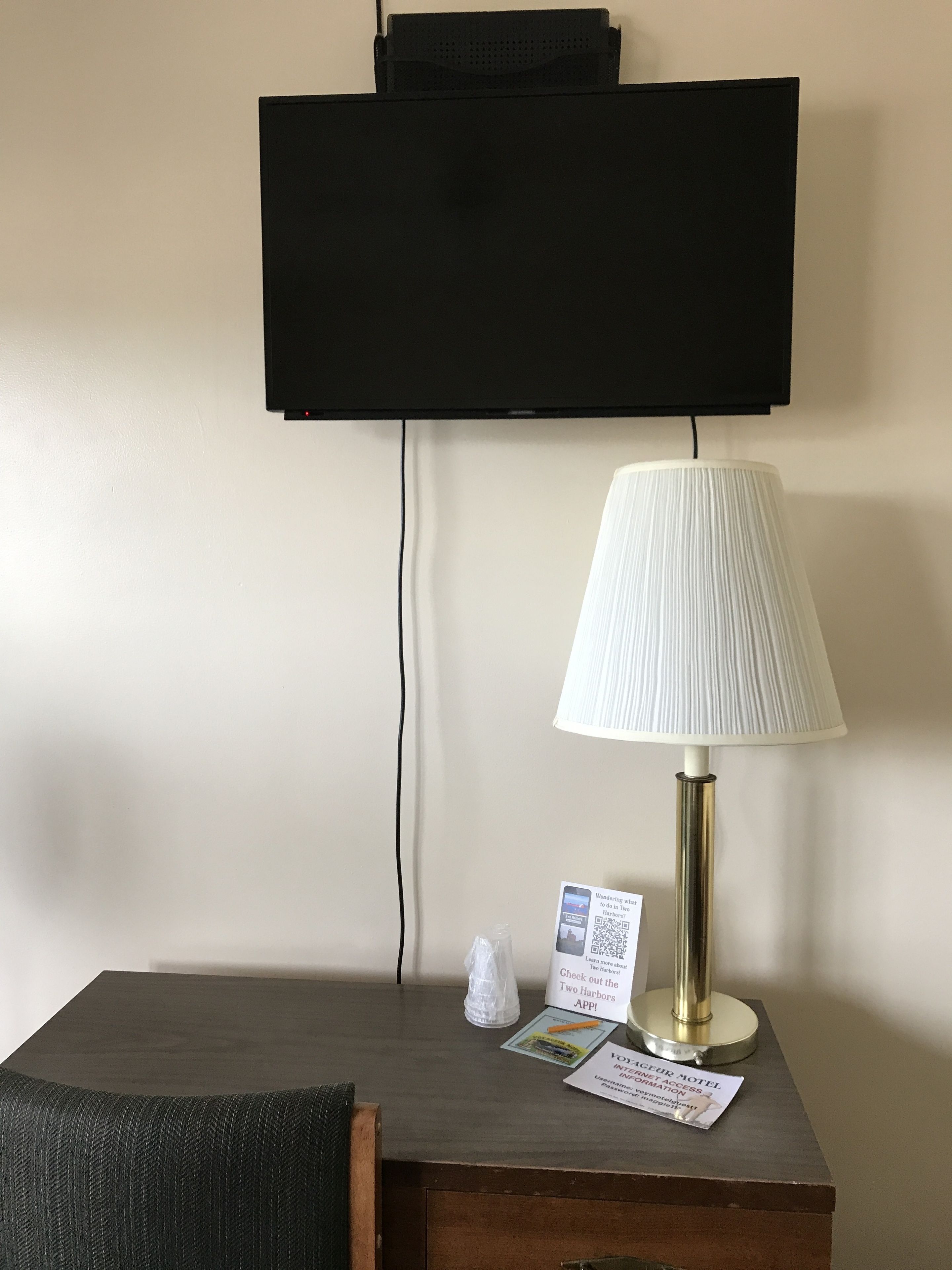 A hotel room with a flat-screen TV mounted on the wall, a table lamp, a small tissue box, and a QR code for the hotel app.