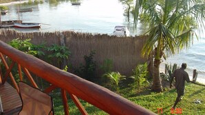 A wooden deck overlooking a calm river with boats, a bamboo fence, and a palm tree. - Casa Chibububo Lodge (Vilanculos)