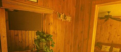 Una habitación de madera con techo de madera, un ventilador de techo, una ventana con persianas, una planta y una puerta.