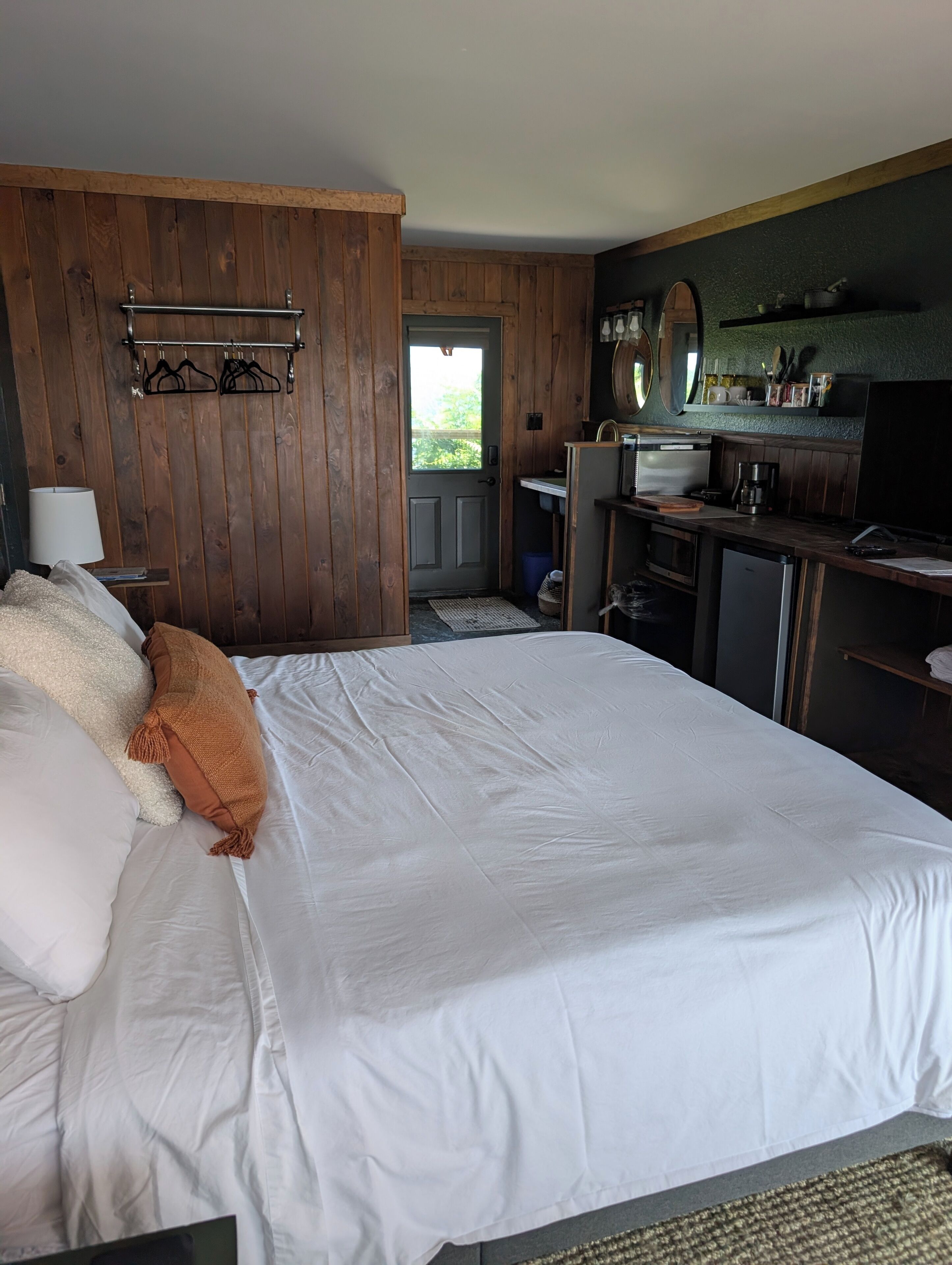 A hotel room with a large bed, a wooden wardrobe, a desk with a chair, a small kitchenette, and a view through a door.