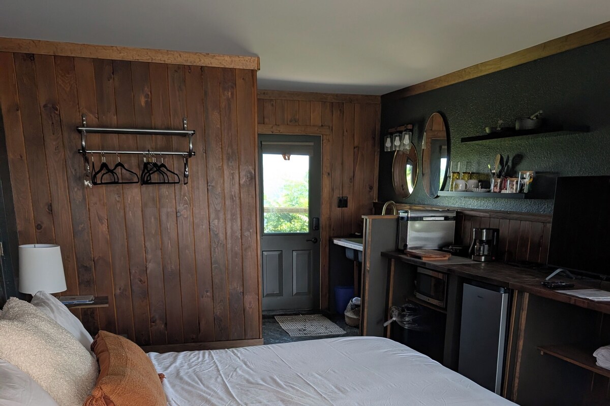 Ein Hotelzimmer mit einem großen Bett, einem Holzschrank, einem Schreibtisch mit Stuhl, einer kleinen Küchenzeile und Blick durch eine Tür.