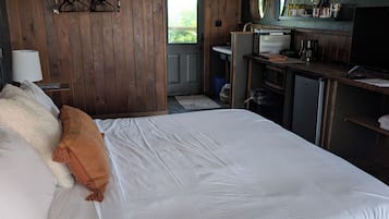 A hotel room with a large bed, a wooden wardrobe, a desk with a chair, a small kitchenette, and a view through a door.