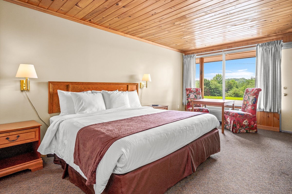 Quarto de hotel com teto de madeira, uma cama grande com roupa de cama branca, duas luminárias de cabeceira, mesa de cabeceira, uma poltrona floral e vista para o verde pela janela.